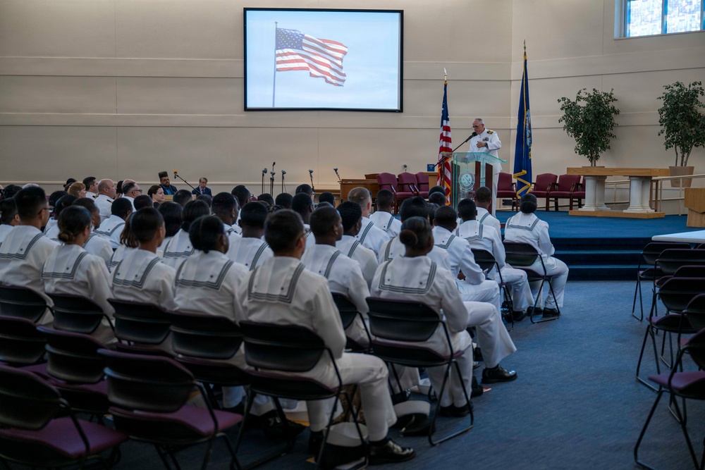 A formal military ceremony is taking place in what appears to be a chapel or auditorium on a naval installation. A senior officer in white dress uniform is speaking at a podium while rows of enlisted sailors in white uniforms are seated facing the front. An American flag is displayed on a large projection screen behind the speaker, with physical flags also flanking the stage.