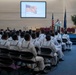 A formal military ceremony is taking place in what appears to be a chapel or auditorium on a naval installation. A senior officer in white dress uniform is speaking at a podium while rows of enlisted sailors in white uniforms are seated facing the front. An American flag is displayed on a large projection screen behind the speaker, with physical flags also flanking the stage.