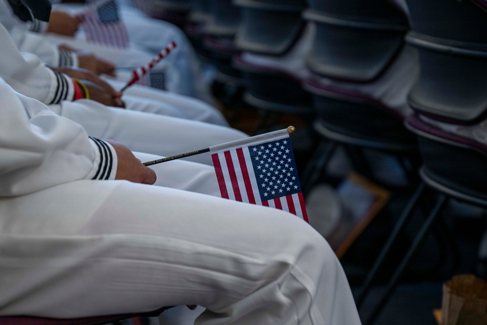The image shows individuals dressed in white military or naval uniforms seated in rows of chairs, each holding small American flags. The flags appear to have text on the sticks, partially readable as 'Citizenship and Immigration Services,' suggesting this is a naturalization ceremony. The close-up composition focuses on the laps and hands of the seated participants.