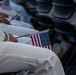 The image shows individuals dressed in white military or naval uniforms seated in rows of chairs, each holding small American flags. The flags appear to have text on the sticks, partially readable as 'Citizenship and Immigration Services,' suggesting this is a naturalization ceremony. The close-up composition focuses on the laps and hands of the seated participants.