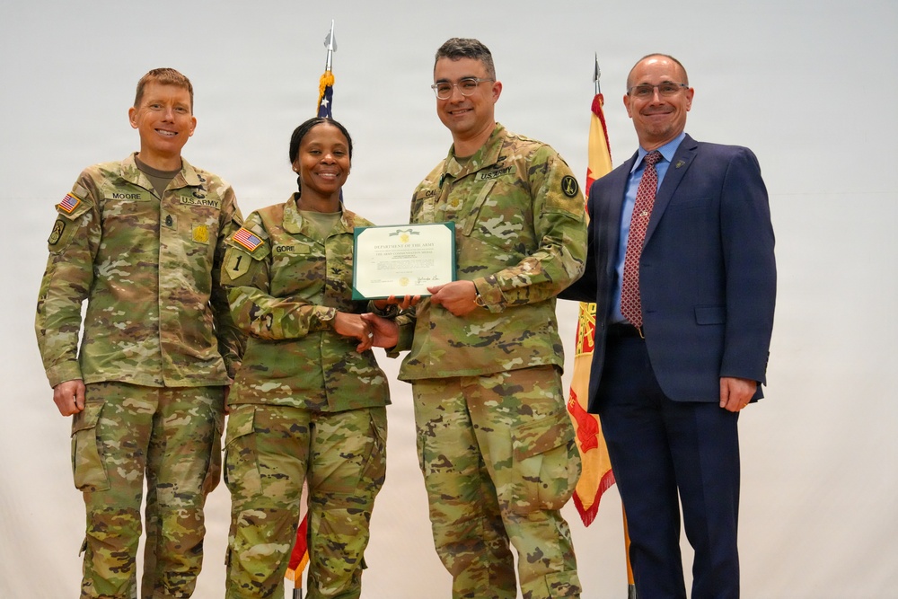 A U.S. Army certificate presentation ceremony featuring four individuals — three in Army OCP (Operational Camouflage Pattern) uniforms and one civilian in a suit. A female soldier identified by nametape as 'GORE' is receiving a Department of the Army certificate from a male soldier identified as 'CAL...' (partially visible), while 'MOORE' stands to the left. The event appears to be a formal military recognition or award ceremony.