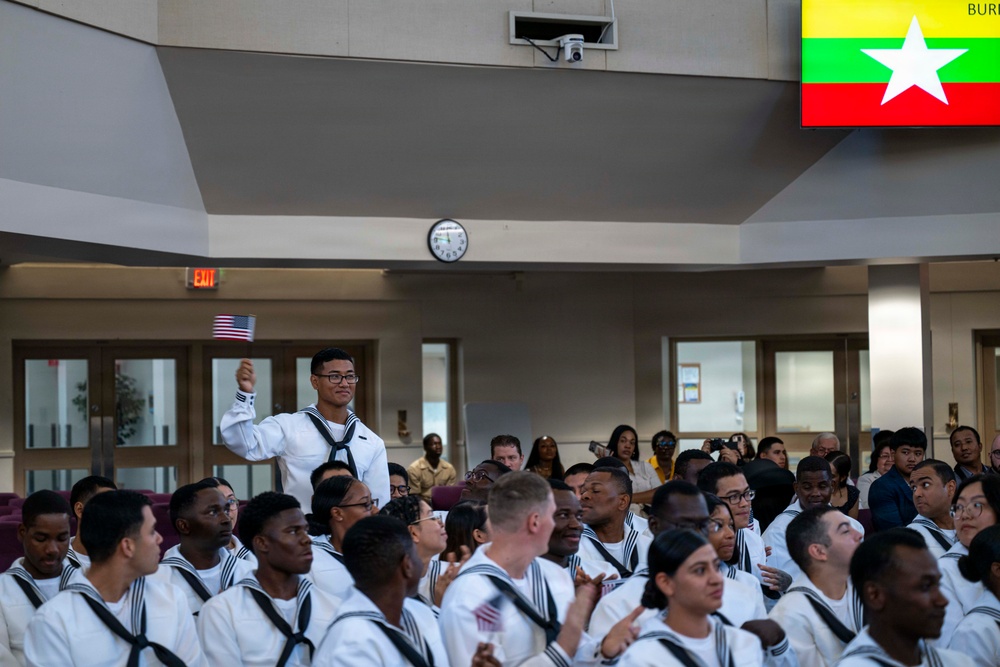 A large group of U.S. Navy recruits in white dress uniforms are seated in an auditorium or ceremony hall. One sailor is standing and raising his fist while holding a small American flag, suggesting a recognition or acknowledgment moment. A Myanmar (Burma) flag is displayed on a screen in the upper right corner, indicating this may be a naturalization or heritage recognition ceremony.