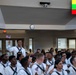 A large group of U.S. Navy recruits in white dress uniforms are seated in an auditorium or ceremony hall. One sailor is standing and raising his fist while holding a small American flag, suggesting a recognition or acknowledgment moment. A Myanmar (Burma) flag is displayed on a screen in the upper right corner, indicating this may be a naturalization or heritage recognition ceremony.