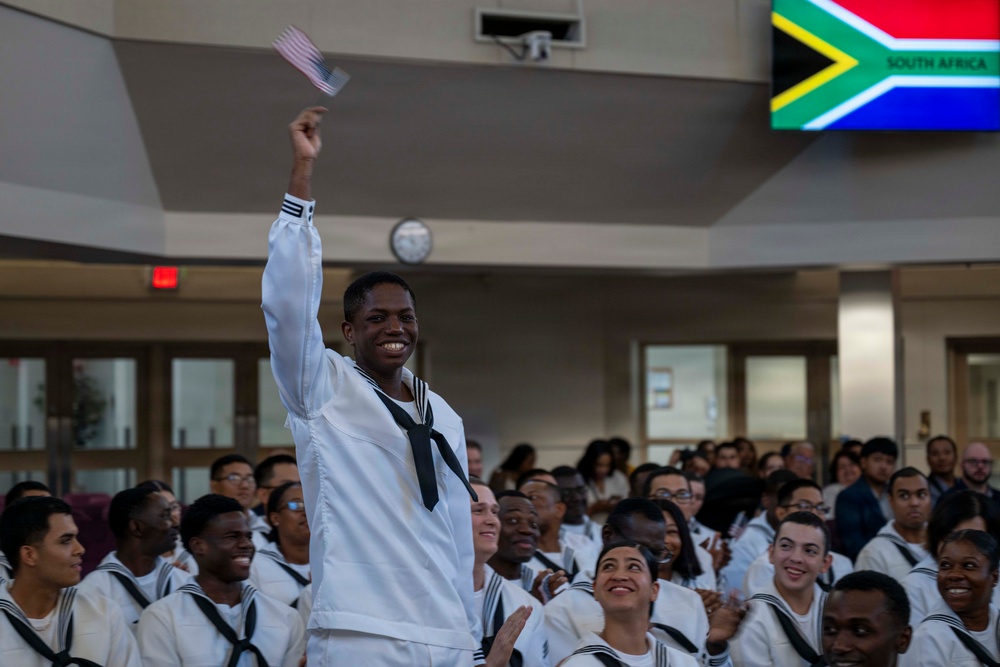 A group of U.S. Navy recruits in white dress uniforms are gathered at what appears to be a naturalization or recognition ceremony. One sailor stands prominently, smiling and waving a small American flag with his arm raised. A screen in the background displays the South African flag with the text 'SOUTH AFRICA,' suggesting this individual or others are being recognized for their country of origin.