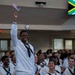A group of U.S. Navy recruits in white dress uniforms are gathered at what appears to be a naturalization or recognition ceremony. One sailor stands prominently, smiling and waving a small American flag with his arm raised. A screen in the background displays the South African flag with the text 'SOUTH AFRICA,' suggesting this individual or others are being recognized for their country of origin.