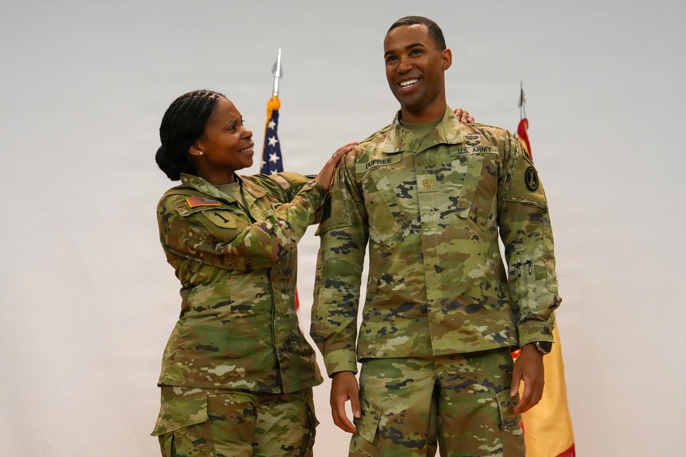 Two U.S. Army soldiers in Operational Camouflage Pattern (OCP) uniforms are posing together during what appears to be a military ceremony or promotion event. The soldier on the right, whose name tape reads 'DUPREE,' is smiling broadly while the soldier on the left places a hand on his shoulder in a congratulatory gesture. An American flag and what appears to be a unit guidon are visible in the background.