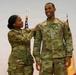 Two U.S. Army soldiers in Operational Camouflage Pattern (OCP) uniforms are posing together during what appears to be a military ceremony or promotion event. The soldier on the right, whose name tape reads 'DUPREE,' is smiling broadly while the soldier on the left places a hand on his shoulder in a congratulatory gesture. An American flag and what appears to be a unit guidon are visible in the background.