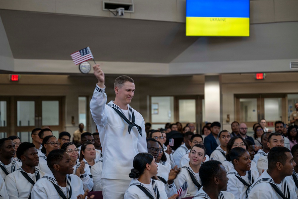 A group of individuals dressed in white U.S. Navy sailor uniforms are gathered in what appears to be a military ceremony or naturalization event. One sailor is standing and waving a small American flag while others remain seated. A screen in the background displays the Ukrainian flag with the word 'UKRAINE' visible.