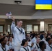 A group of individuals dressed in white U.S. Navy sailor uniforms are gathered in what appears to be a military ceremony or naturalization event. One sailor is standing and waving a small American flag while others remain seated. A screen in the background displays the Ukrainian flag with the word 'UKRAINE' visible.