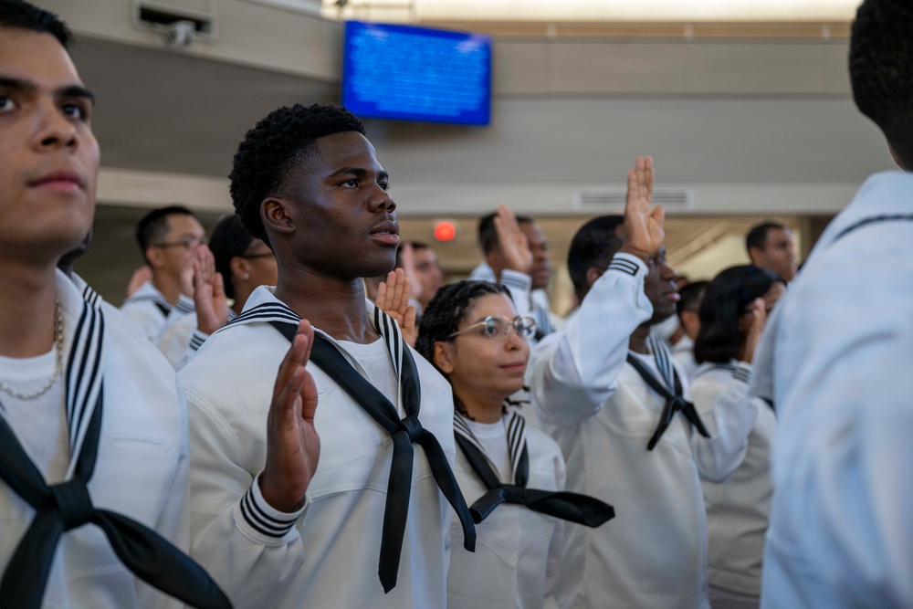 A group of individuals dressed in white U.S. Navy enlisted uniforms with black neckerchiefs are raising their right hands, appearing to take an oath or pledge. The scene appears to be an enlistment or re-enlistment ceremony taking place indoors, likely at a military processing or recruitment facility. A blue display screen is visible in the background.