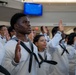 A group of individuals dressed in white U.S. Navy enlisted uniforms with black neckerchiefs are raising their right hands, appearing to take an oath or pledge. The scene appears to be an enlistment or re-enlistment ceremony taking place indoors, likely at a military processing or recruitment facility. A blue display screen is visible in the background.