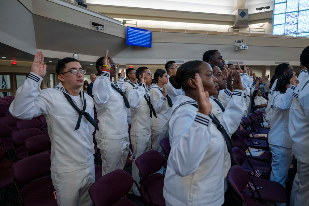 A group of U.S. Navy recruits dressed in white dress uniforms are raising their right hands, appearing to take an oath of enlistment or re-enlistment during a formal ceremony. The event is taking place in a large auditorium or chapel-like venue with purple/maroon seating. Small American flags are visible among the participants.