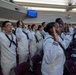 A group of U.S. Navy recruits dressed in white dress uniforms are raising their right hands, appearing to take an oath of enlistment or re-enlistment during a formal ceremony. The event is taking place in a large auditorium or chapel-like venue with purple/maroon seating. Small American flags are visible among the participants.