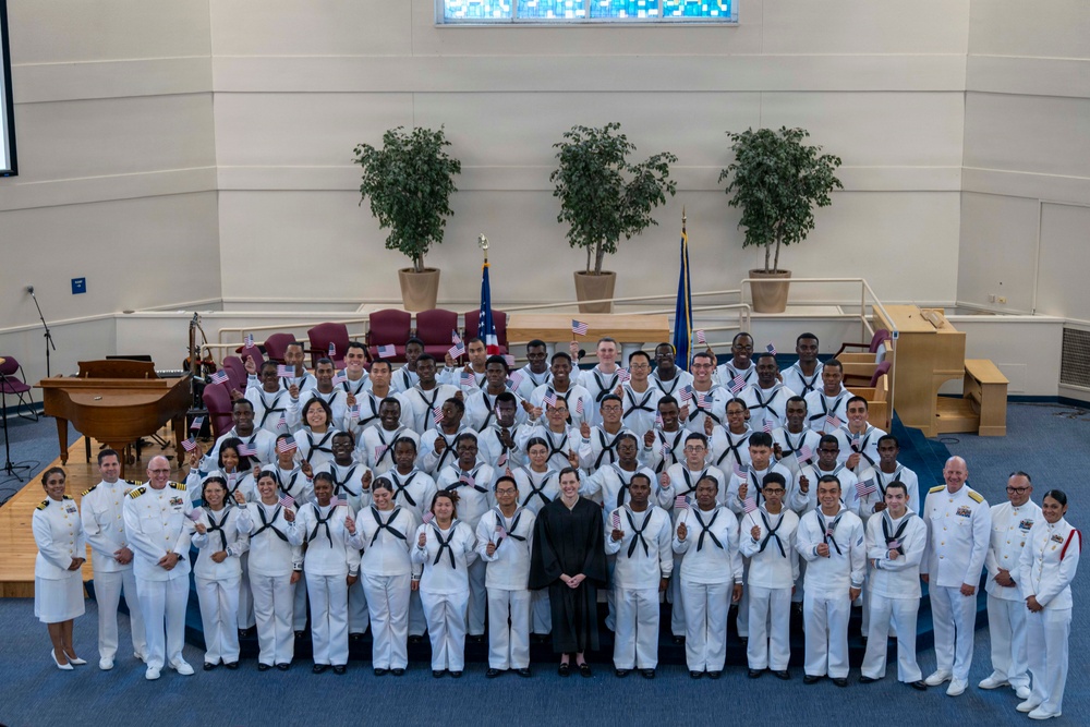 A large group photograph of U.S. Navy personnel in white dress uniforms, gathered in what appears to be a chapel or auditorium on a military base. Many of the sailors are holding small American flags, and a person in judicial robes stands at the center, suggesting this is a naturalization ceremony. Senior officers in white service dress uniforms flank the group on both sides.