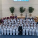 A large group photograph of U.S. Navy personnel in white dress uniforms, gathered in what appears to be a chapel or auditorium on a military base. Many of the sailors are holding small American flags, and a person in judicial robes stands at the center, suggesting this is a naturalization ceremony. Senior officers in white service dress uniforms flank the group on both sides.