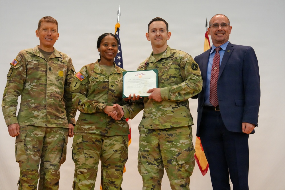 Four individuals pose for a formal military award ceremony photograph. Two U.S. Army soldiers in the center are shaking hands while jointly holding a certificate or commendation document. A civilian in a blue suit stands to the right, while another uniformed soldier stands to the left.