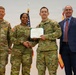 Four individuals pose for a formal military award ceremony photograph. Two U.S. Army soldiers in the center are shaking hands while jointly holding a certificate or commendation document. A civilian in a blue suit stands to the right, while another uniformed soldier stands to the left.