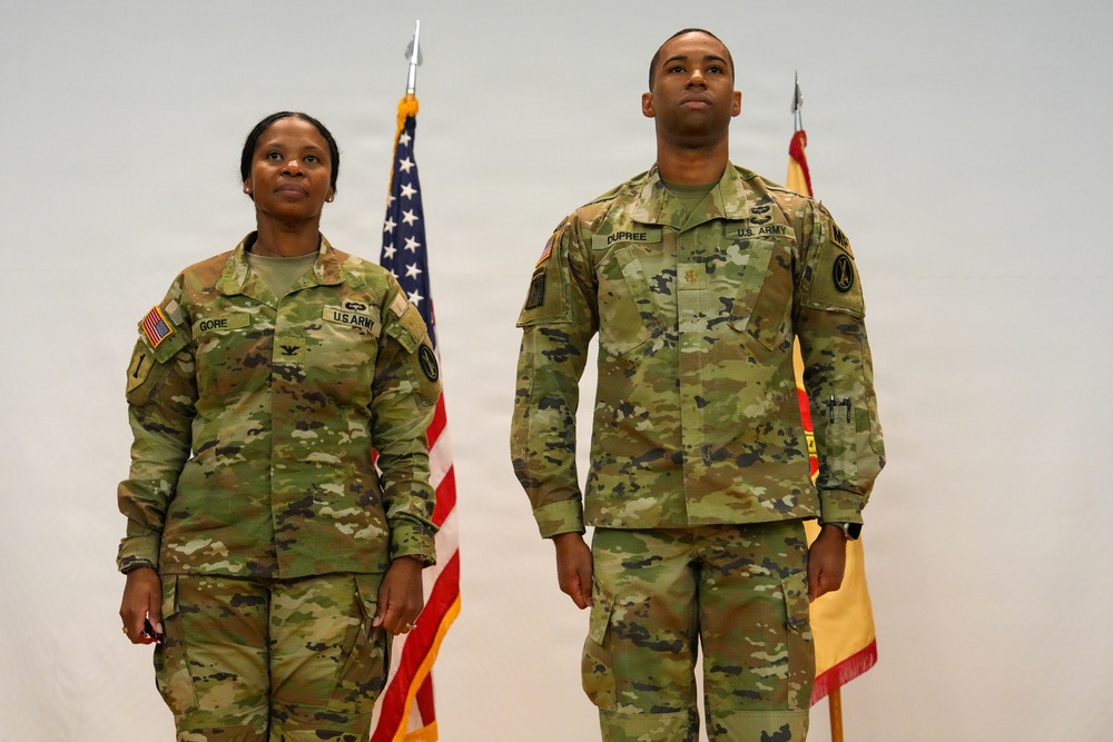 Two U.S. Army soldiers in OCP (Operational Camouflage Pattern) uniforms stand at attention during what appears to be a formal military ceremony. The soldier on the left has the name tape 'GORE' and displays a Colonel's eagle insignia, while the soldier on the right bears the name tape 'DUPREE' and wears U.S. Army insignia. An American flag and a unit guidon flag are visible in the background.