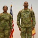 Two U.S. Army soldiers in OCP (Operational Camouflage Pattern) uniforms stand at attention during what appears to be a formal military ceremony. The soldier on the left has the name tape 'GORE' and displays a Colonel's eagle insignia, while the soldier on the right bears the name tape 'DUPREE' and wears U.S. Army insignia. An American flag and a unit guidon flag are visible in the background.