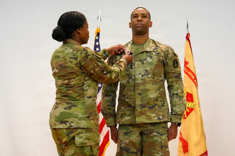 A U.S. Army military ceremony in which a female soldier is pinning a medal or award onto a male soldier's uniform. Both individuals are wearing OCP (Operational Camouflage Pattern) uniforms. An American flag and a unit guidon flag are visible in the background.