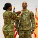 A U.S. Army military ceremony in which a female soldier is pinning a medal or award onto a male soldier's uniform. Both individuals are wearing OCP (Operational Camouflage Pattern) uniforms. An American flag and a unit guidon flag are visible in the background.