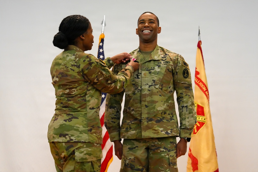 Two U.S. Army soldiers in Operational Camouflage Pattern (OCP) uniforms are participating in a military pinning ceremony. A female soldier is pinning a decoration or insignia onto the chest of a male soldier, who is smiling broadly. The ceremony takes place in front of a U.S. flag and what appears to be a unit guidon or organizational flag.