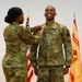 Two U.S. Army soldiers in Operational Camouflage Pattern (OCP) uniforms are participating in a military pinning ceremony. A female soldier is pinning a decoration or insignia onto the chest of a male soldier, who is smiling broadly. The ceremony takes place in front of a U.S. flag and what appears to be a unit guidon or organizational flag.