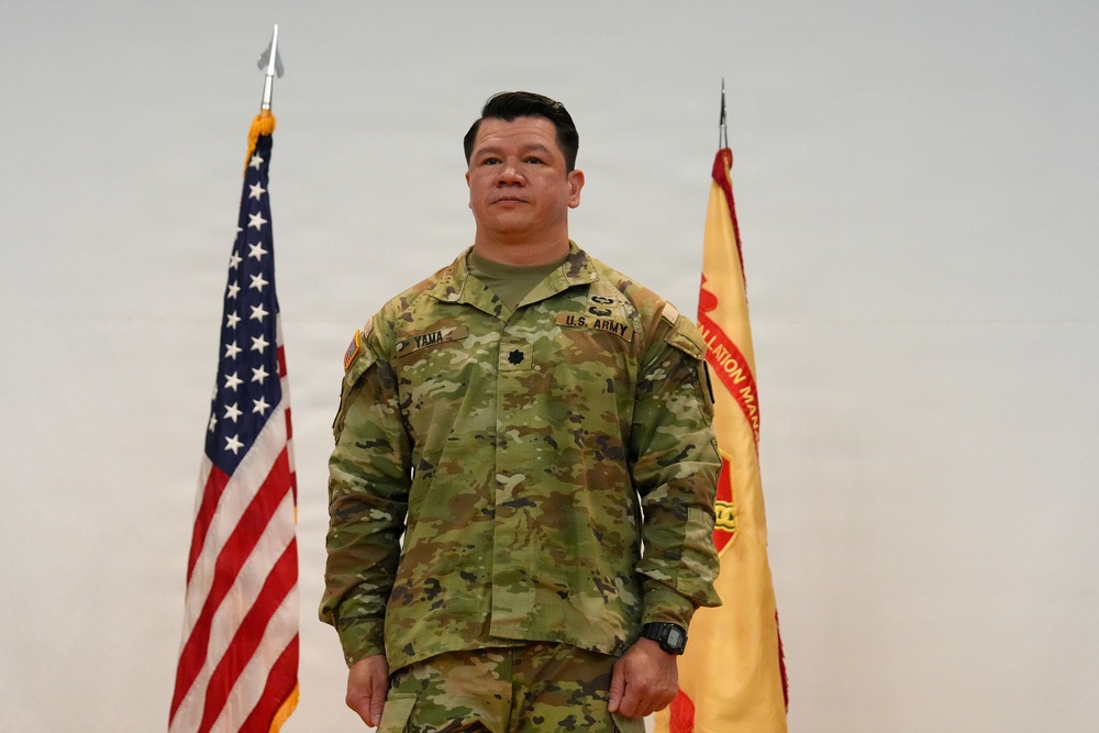 A U.S. Army officer in Operational Camouflage Pattern (OCP) uniform stands at attention between an American flag and what appears to be an Installation Management Command flag. The name tape reads 'YANA' and a Lieutenant Colonel rank insignia (oak leaf) is visible on the chest. This appears to be an official military portrait or ceremony photograph.