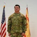 A U.S. Army officer in Operational Camouflage Pattern (OCP) uniform stands at attention between an American flag and what appears to be an Installation Management Command flag. The name tape reads 'YANA' and a Lieutenant Colonel rank insignia (oak leaf) is visible on the chest. This appears to be an official military portrait or ceremony photograph.