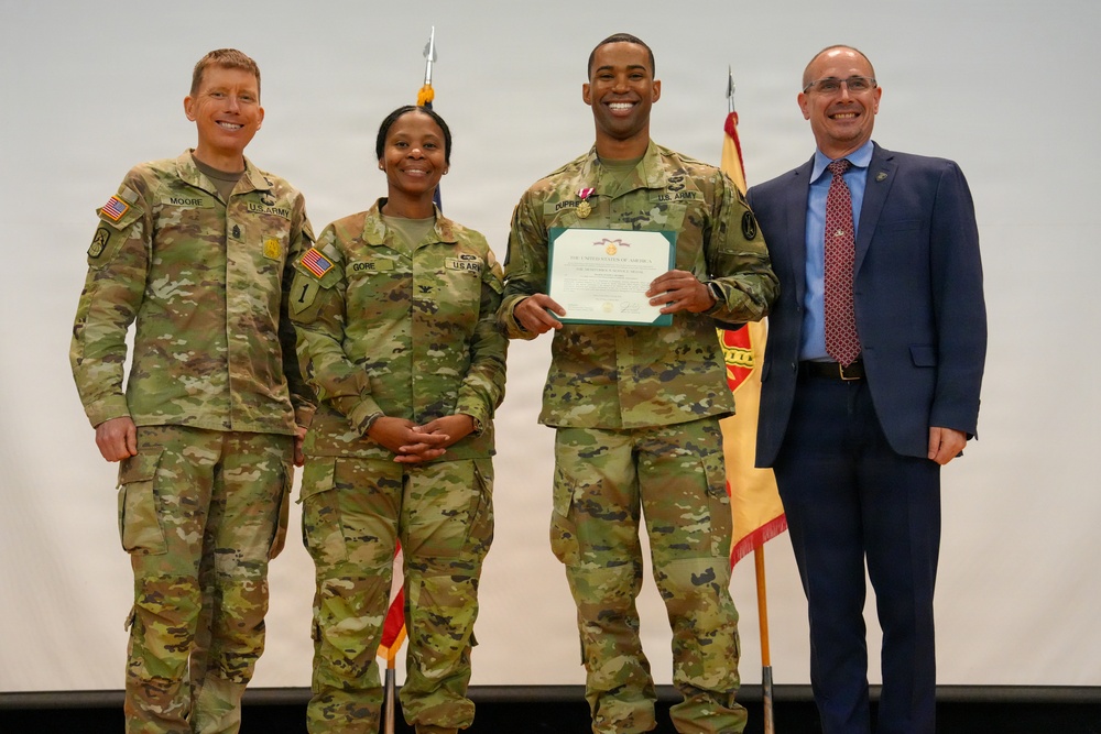 A military award ceremony featuring four individuals posing for a formal photo. Three are wearing U.S. Army OCP (Operational Camouflage Pattern) uniforms, while one civilian in a blue suit stands to the right. The central soldier is holding a certificate and wearing a medal, indicating a formal recognition or commendation event.