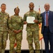 A military award ceremony featuring four individuals posing for a formal photo. Three are wearing U.S. Army OCP (Operational Camouflage Pattern) uniforms, while one civilian in a blue suit stands to the right. The central soldier is holding a certificate and wearing a medal, indicating a formal recognition or commendation event.