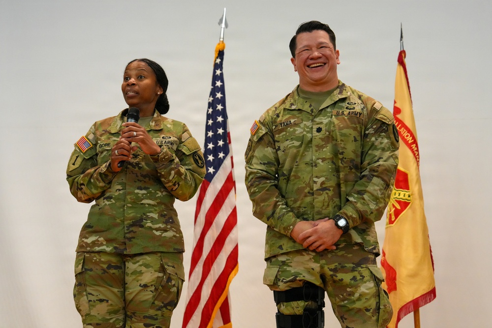 Two U.S. Army soldiers in OCP (Operational Camouflage Pattern) uniforms stand before an audience at what appears to be a military ceremony or event. The soldier on the left, identified by name tape as 'GORE,' is speaking into a microphone, while the soldier on the right, identified as 'YAMA,' is laughing. An American flag and a unit guidon flag are visible in the background.