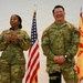 Two U.S. Army soldiers in OCP (Operational Camouflage Pattern) uniforms stand before an audience at what appears to be a military ceremony or event. The soldier on the left, identified by name tape as 'GORE,' is speaking into a microphone, while the soldier on the right, identified as 'YAMA,' is laughing. An American flag and a unit guidon flag are visible in the background.
