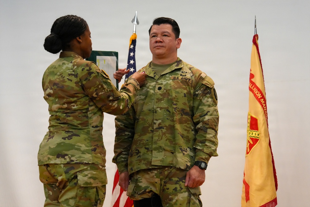A U.S. Army promotion or award ceremony is taking place, with one soldier pinning an insignia or medal onto another soldier's uniform. The recipient is wearing U.S. Army OCP (Operational Camouflage Pattern) uniform with a rank insignia visible on the chest. An American flag and what appears to be an Installation Management Command flag are visible in the background.