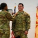 A U.S. Army promotion or award ceremony is taking place, with one soldier pinning an insignia or medal onto another soldier's uniform. The recipient is wearing U.S. Army OCP (Operational Camouflage Pattern) uniform with a rank insignia visible on the chest. An American flag and what appears to be an Installation Management Command flag are visible in the background.