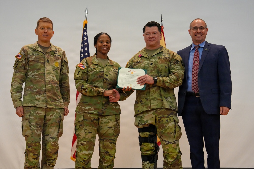 A U.S. Army award ceremony showing four individuals posing for a photo. Two soldiers are jointly holding a certificate/award document, while a third soldier and a civilian in a suit stand alongside them. The setting appears to be an indoor military ceremony with American and unit flags visible in the background.