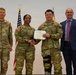 A U.S. Army award ceremony showing four individuals posing for a photo. Two soldiers are jointly holding a certificate/award document, while a third soldier and a civilian in a suit stand alongside them. The setting appears to be an indoor military ceremony with American and unit flags visible in the background.