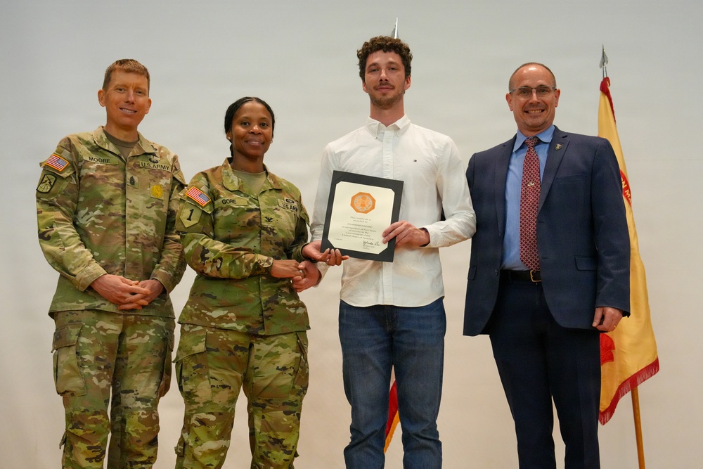 Four individuals pose for a formal photo during what appears to be a military award or recognition ceremony. Two U.S. Army soldiers in OCP (Operational Camouflage Pattern) uniforms flank a civilian male holding a certificate/award, with a fourth person in civilian business attire standing to the right. A military unit guidon or organizational flag is visible in the background on the right side.