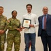 Four individuals pose for a formal photo during what appears to be a military award or recognition ceremony. Two U.S. Army soldiers in OCP (Operational Camouflage Pattern) uniforms flank a civilian male holding a certificate/award, with a fourth person in civilian business attire standing to the right. A military unit guidon or organizational flag is visible in the background on the right side.