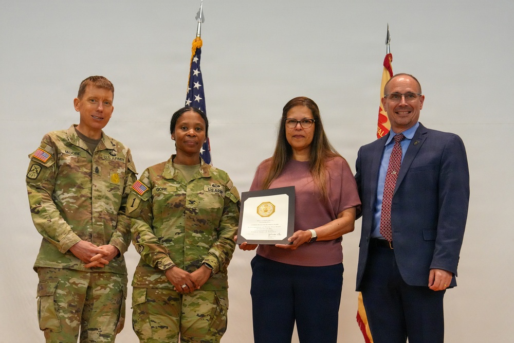 Four individuals pose for a formal photograph during what appears to be a military award or recognition ceremony. Two U.S. Army soldiers in OCP (Operational Camouflage Pattern) uniforms flank a civilian woman holding a framed certificate or award, alongside a man in a business suit. An American flag and a red military organizational flag are visible in the background.