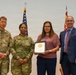 Four individuals pose for a formal photograph during what appears to be a military award or recognition ceremony. Two U.S. Army soldiers in OCP (Operational Camouflage Pattern) uniforms flank a civilian woman holding a framed certificate or award, alongside a man in a business suit. An American flag and a red military organizational flag are visible in the background.