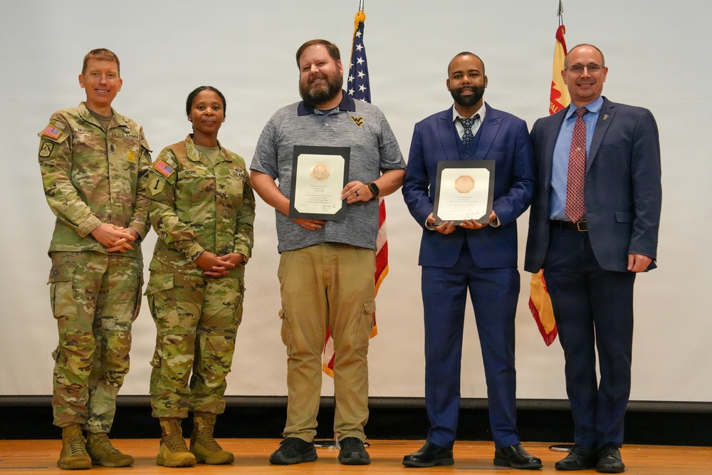 Five individuals pose for a formal group photo at what appears to be a military awards ceremony. Two U.S. Army soldiers in camouflage uniforms stand alongside two civilians holding framed award certificates and one civilian in a suit. An American flag and what appears to be a military unit flag are visible in the background.