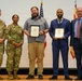 Five individuals pose for a formal group photo at what appears to be a military awards ceremony. Two U.S. Army soldiers in camouflage uniforms stand alongside two civilians holding framed award certificates and one civilian in a suit. An American flag and what appears to be a military unit flag are visible in the background.