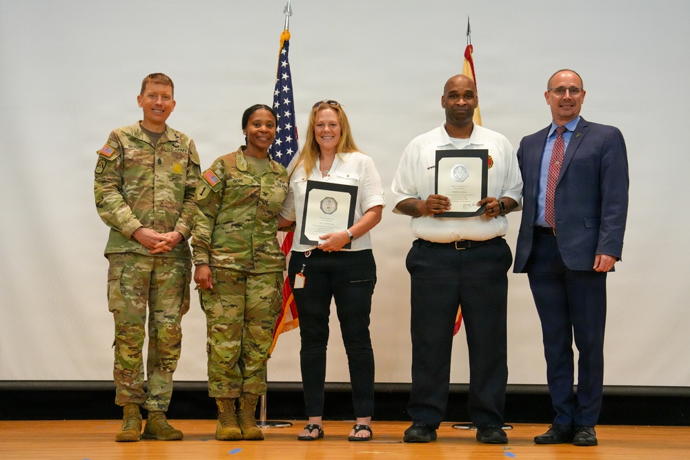 Five individuals pose for a formal group photo during what appears to be a military awards or recognition ceremony. Two uniformed U.S. Army soldiers stand alongside two civilians holding framed certificates or plaques, with a man in a business suit on the far right. Two American flags are visible in the background against a white projection screen.