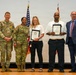 Five individuals pose for a formal group photo during what appears to be a military awards or recognition ceremony. Two uniformed U.S. Army soldiers stand alongside two civilians holding framed certificates or plaques, with a man in a business suit on the far right. Two American flags are visible in the background against a white projection screen.