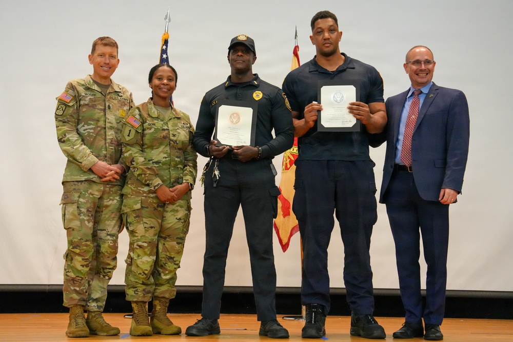Five individuals are posed together on a stage during what appears to be a military or government awards ceremony. Two U.S. Army soldiers in camouflage uniforms stand alongside two security personnel in dark uniforms holding official certificates, and one civilian in a blue suit. Flags including the U.S. flag are visible in the background.
