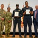 Five individuals are posed together on a stage during what appears to be a military or government awards ceremony. Two U.S. Army soldiers in camouflage uniforms stand alongside two security personnel in dark uniforms holding official certificates, and one civilian in a blue suit. Flags including the U.S. flag are visible in the background.