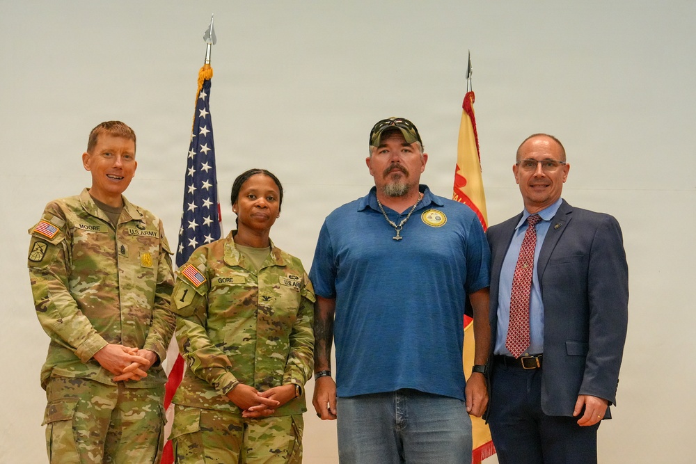 Four individuals pose for a formal group photo in front of an American flag and what appears to be a New Mexico state flag. Two are wearing U.S. Army combat uniforms (OCP/multicam), one is in civilian casual attire, and one is in business professional clothing. The setting appears to be an official military or government ceremony or event.