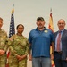 Four individuals pose for a formal group photo in front of an American flag and what appears to be a New Mexico state flag. Two are wearing U.S. Army combat uniforms (OCP/multicam), one is in civilian casual attire, and one is in business professional clothing. The setting appears to be an official military or government ceremony or event.