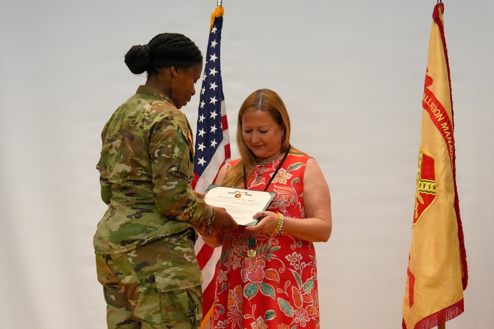 A U.S. Army soldier in OCP (Operational Camouflage Pattern) uniform is presenting a certificate or award to a civilian woman wearing a red floral dress. The ceremony appears to be a formal military recognition event, with both individuals looking down at the document being exchanged. Two flags are visible in the background — the U.S. national flag and what appears to be a unit or installation management flag.